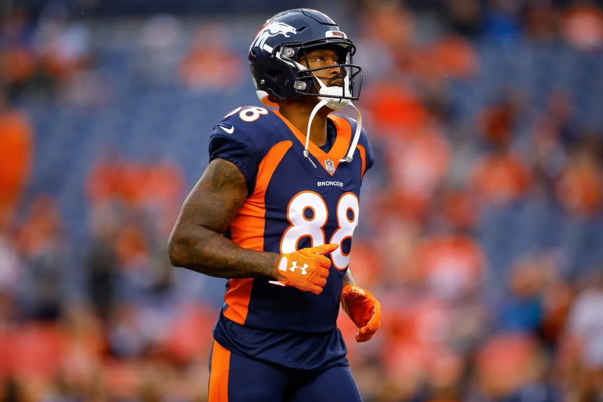 A football player in a navy and orange Denver Broncos uniform with the number 88, wearing a helmet and running on a field with a blurred stadium crowd in the background.